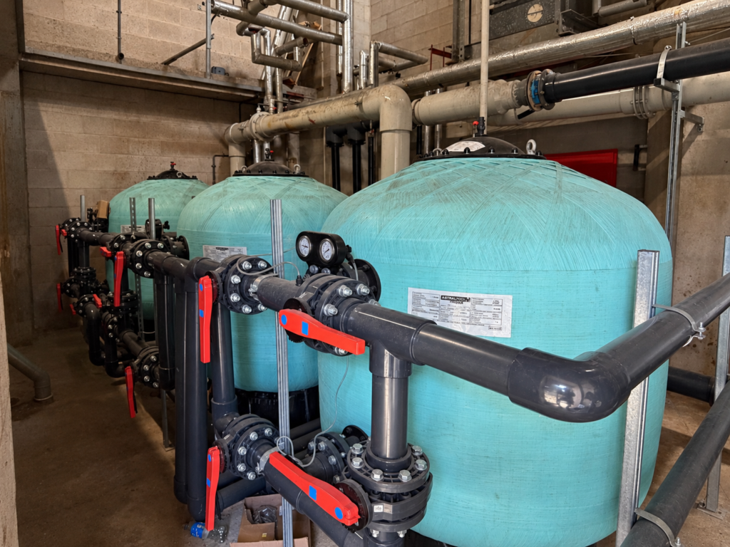 Three commercial pool filters with grey circulation pipework and red valve handles